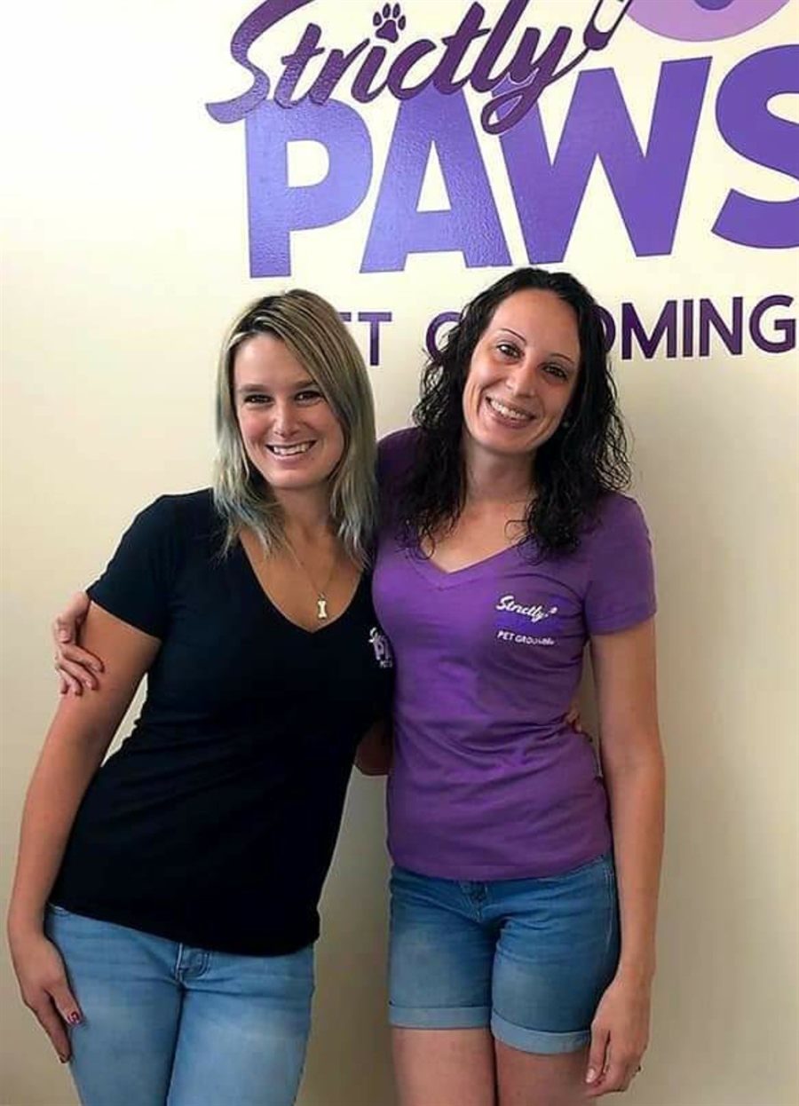 Josie, owner and groomer, with Jackie, the salon's bather, smiling in front of the purple Strictly Paws logo wall